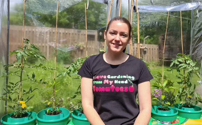 Sahara sits inside a greenhouse with tomato plants, wearing a t-shirt that reads: I love gardening from my head tomatoes